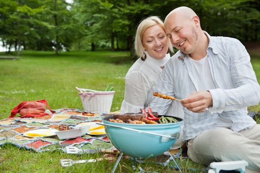 Happy mature couple cooking meat on portable barbecue at an outdoor picnic in forest park