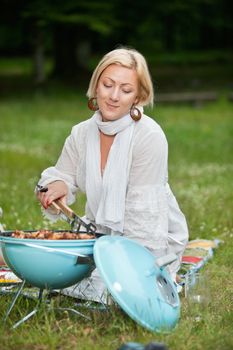 Beautiful mature woman in casual wear cooking food on a portable barbecue on a weekend outing