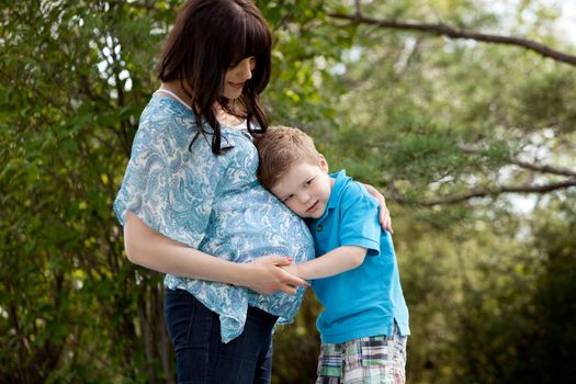 Young son listening to belly of pregnant outdoors in park