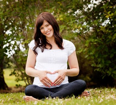 Portrait of a happy healthy pregnant woman in park making heart sign on belly