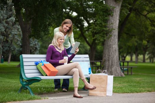Beautiful young female friends with shopping bags using tablet PC on park bench