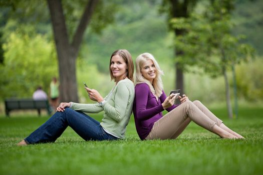 Side view portrait of happy young female friends with cellphones sitting back to back in park