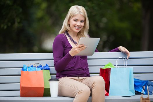 Portrait of pretty young woman with shopping bags using tablet PC on park bench