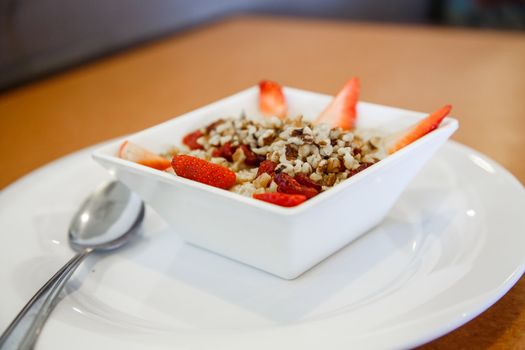 Steel-cut oatmeal in white bowl on white plate with strawberries, dried cranberries and walnuts