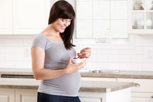 Happy pregnant woman in kitchen eating a healthy snack of strawberries and granola