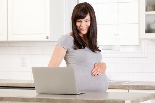 Happy pregnant woman at home in kitchen using laptop computer