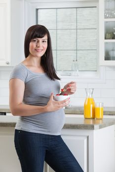 Portrait of happy pregnant woman eating healthy bowl of fruit in kitchen