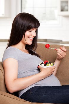 Happy smiling pregnant woman sitting on sofa eating fresh fruit