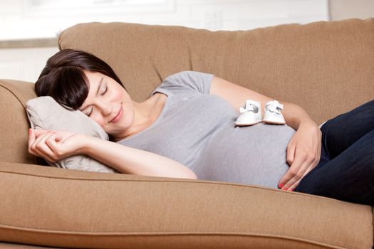 Content pregnant mother resting on sofa in living room with baby shoes on belly