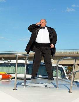 Overweight man in tuxedo standing on the deck of a luxury pleasure boat with glass red wine