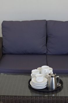 A tray with empty coffee cups and a full coffee pot and sugar bowl placed on a rattan table with a glass top, in front of a rattan sofa with blue cushions