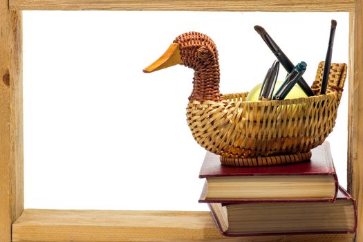 Wicker basket with a ball-point pens and brushes and Some books with dark red hardcover and green apple in a wooden frame on white background