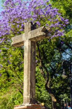 A stone cross in Mexico City with purple flowers and trees in the background