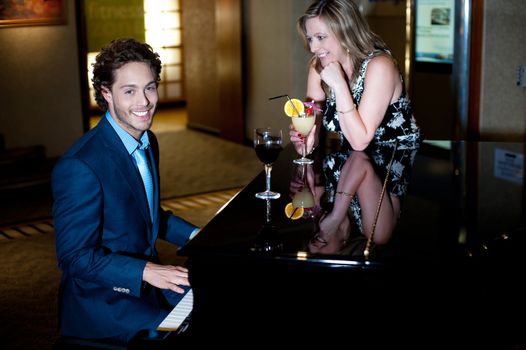 Attractive young man playing piano and entertaining his companion holding cocktail. Love couple