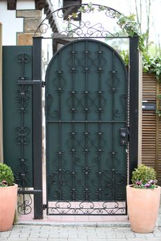 Ornate green metal entry gate for pedestrians with fancy wrought iron pattern detail set into a high wall between two potted plants with the facade of a private residence visible behind