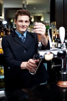 Handsome mixologist putting ice into tall glass. Bar decorated with bottles in the background