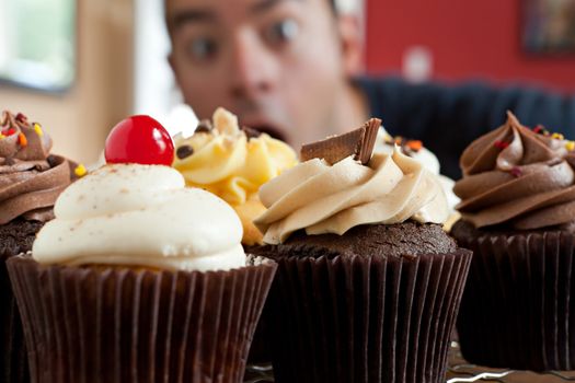 Close up of some decadent gourmet cupcakes frosted with a variety of frosting flavors.  Shallow depth of field with the face of a hungry man lurking in the background.