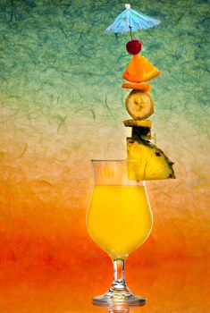 An orange drink with a tower of citrus fruits including a blue umbrella against an abstract sunset colored background.