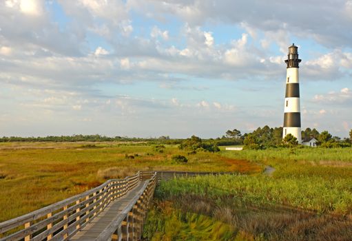 Walkway leading to the Bodie Island lighthouse over marshes of the Cape Hatteras National Seashore against white clouds and a blue morning sky