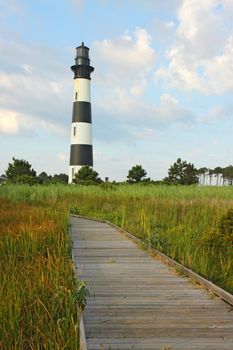 Walkway leading to the Bodie Island lighthouse over marshes of the Cape Hatteras National Seashore against white clouds and a blue morning sky vertical