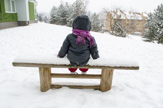 child with a dark gray jacket and purple scarf sitting on a bench abundant snow in the distance snow pines top