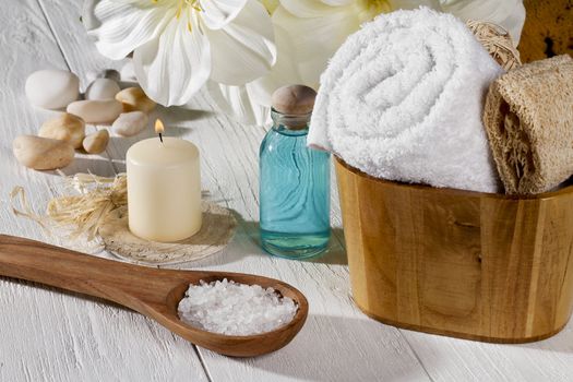 A set of spa products with spa salt on a spoon, towel, oil and a lighted candle on a wooden table