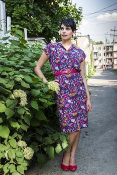 Young fashionable woman wearing a multi colored flower pattern dress near a flower bush in an urban alley