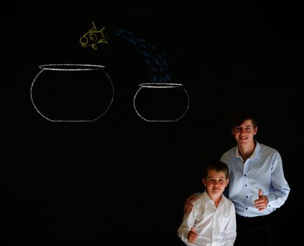 Thumbs up boy dressed up as business man with teacher man and fish jumping from small bowl to big bowl on blackboard background