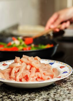 Closeup of raw chicken dish and female cooking vegetables in a black pan in the background