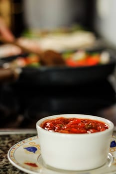 Closeup of bowl of hot sauce prepared for a mexican food and female cooking in a black pan in the background