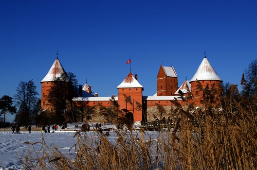 People tourist recreate near ancient Trakai castle fortification covered with snow near frozen lake and reeds moving in wind.