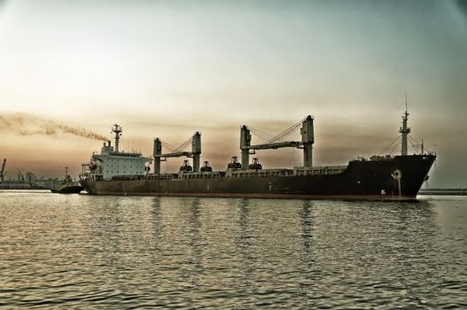 The stylized semi-antique photo. The ore carrier heaves out of the harbor on a tow, the photo is taken at sunset.