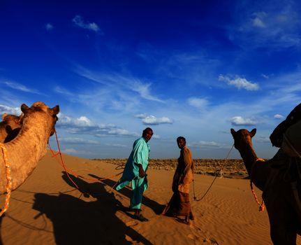 Rajasthan travel background - two indian cameleers (camel drivers) with camels in dunes of Thar desert. Jaisalmer, Rajasthan, India