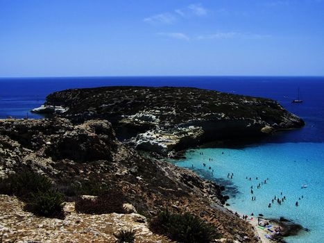 This is the magnificent island of rabbits, in Lampedusa. The water is crystal clear and the sand is white. The rocks are silhouetted against the blue sea and the sky is clear. This area is protected reserves.