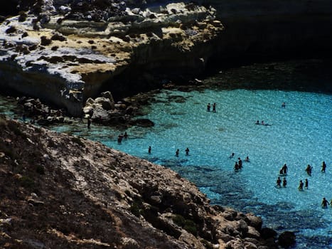 This is the magnificent island of rabbits, in Lampedusa. The water is crystal clear and the sand is white. The rocks are silhouetted against the blue sea and the sky is clear. This area is protected reserves.