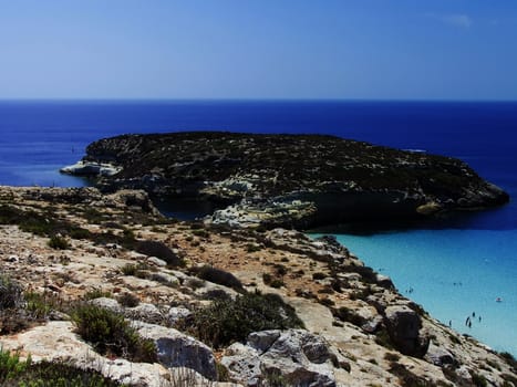This is the magnificent island of rabbits, in Lampedusa. The water is crystal clear and the sand is white. The rocks are silhouetted against the blue sea and the sky is clear. This area is protected reserves.
