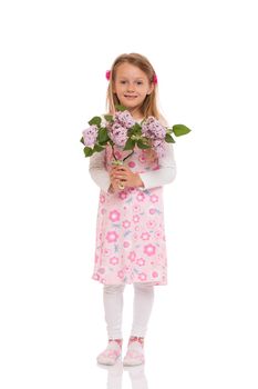 Smiling little girl with long hair wearing summer dress standing and holding lilac flowers. Isolated on white background.