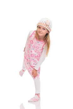 Full length portrait of a funny little girl with long hair wearing summer dress, knit cap and balancing at one leg. Isolated on white background.