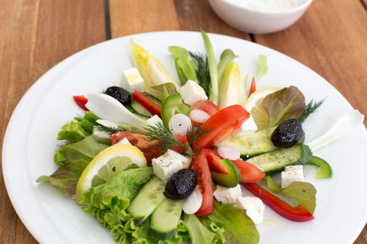 Greek salad with feta, tomatoes, cucumber, peppers and black olives. With bowl of sauce on a wooden table.