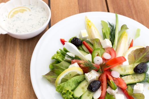 Greek salad with feta, tomatoes, cucumber, peppers and black olives. With bowl of sauce on a wooden table.