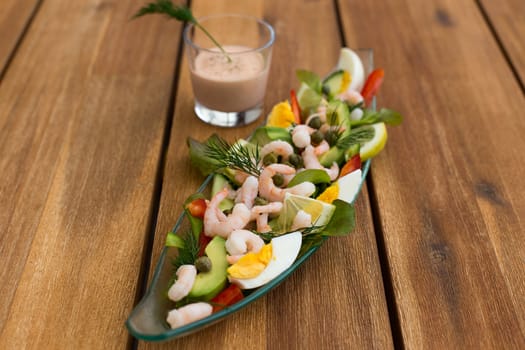 Shrimp cocktail with avocado, eggs, lemon and glass of sauce on a wooden table.