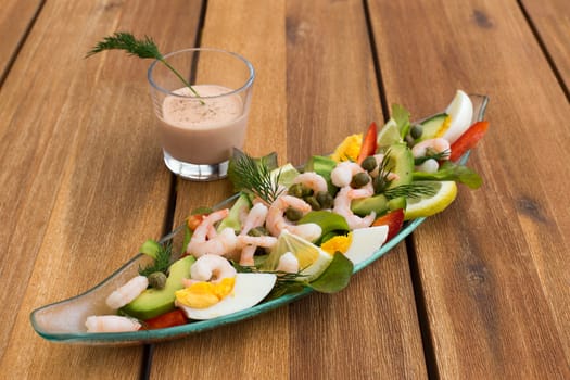 Shrimp cocktail with avocado, eggs, lemon and glass of sauce on a wooden table.