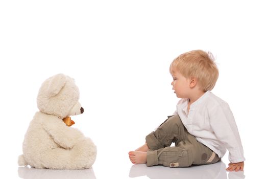 Little boy with blond hair sitting on the floor and looking at her teddy bear. Isolated on white background.
