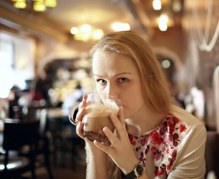 Portrait of girl enjoying coffee latte in cafe and looking to the photographer. Beautiful vintage interior in blur with natural sunlight.