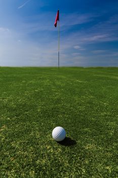 Close up of a ball on a green with a flag in the distance against a blue sky