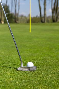Close up of a golf ball and a putter on a green with a flag in the distance