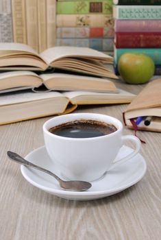 A cup of coffee on the table against the background of an open book with a notebook and a stack of books