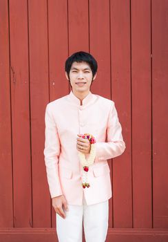 Asian Thai man/groom in Thai wedding suit is holding flowered garland.