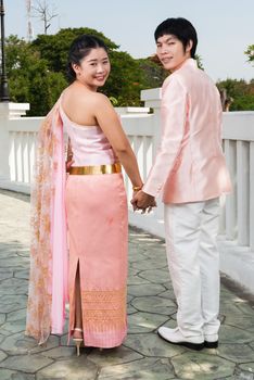 Asian Thai groom is holding his bride's hand beside white bridge.