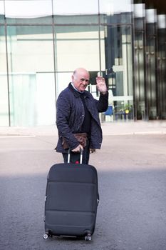 Elderly man waving goodbye as he makes his way to the departure terminal with his suitcase at the end of his holiday